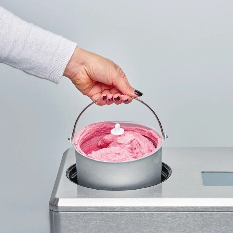 Ręka trzyma wiadro z różowymi lodami. Wiadro jest w srebrnej maszynie. Tło jest gładko białe.