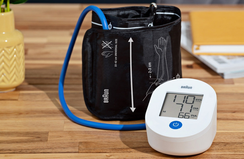 Ciśnieniomierz Braun z mankietem na ramię i cyfrowym wyświetlaczem. Jest na drewnianej powierzchni. Po lewej stronie znajduje się żółty wazon.