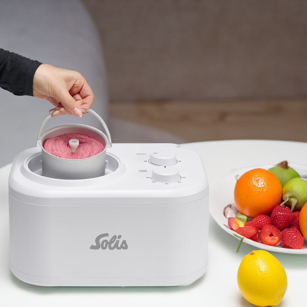 Osoba trzyma różowy pojemnik na lody z białej maszyny Solis. W pobliżu znajduje się talerz owoców, w tym pomarańcze i truskawki.