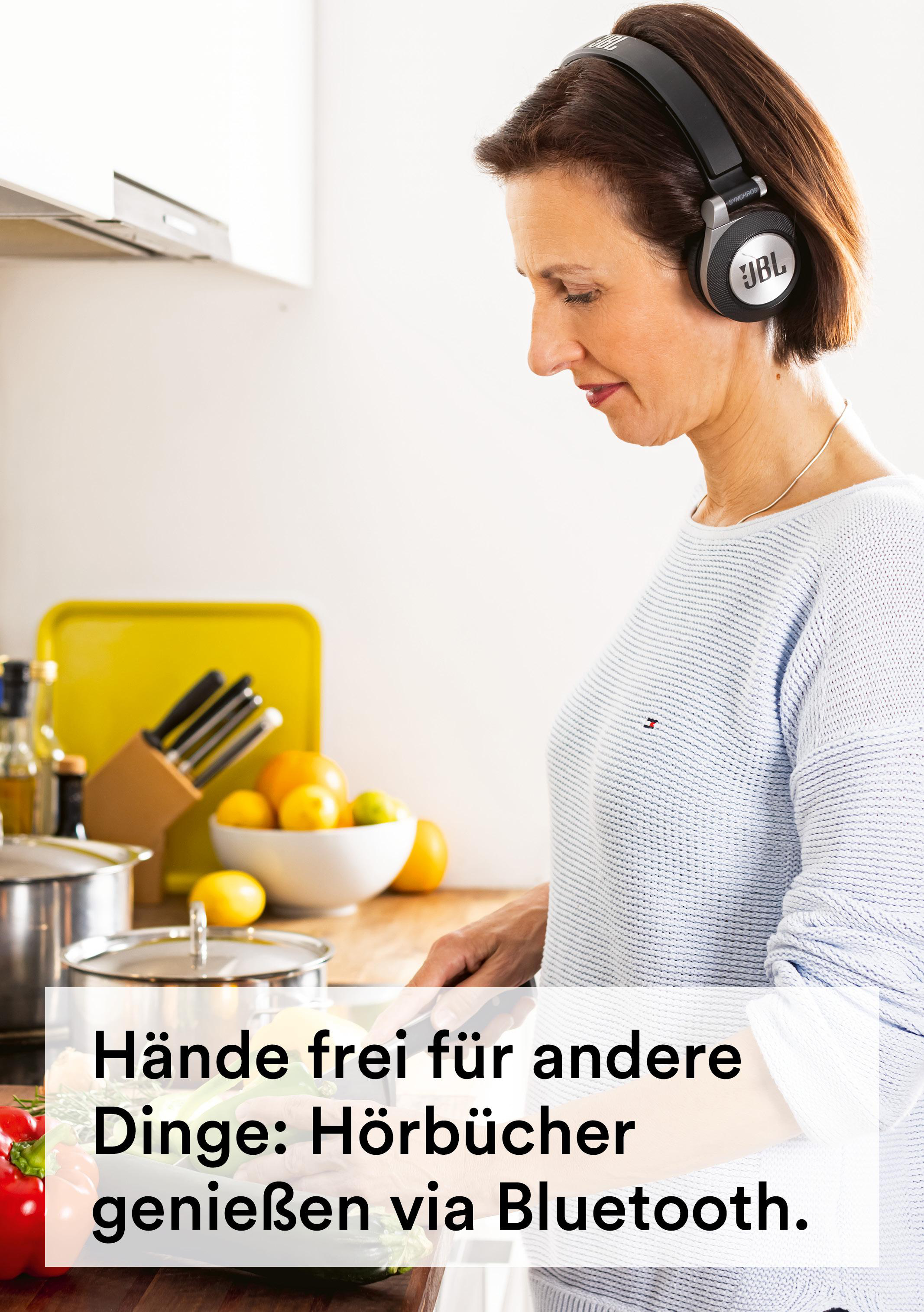Frau in der Küche mit Kopfhörern, schneidet Gemüse. Gelbes Tablett, Schüssel mit Zitronen und Töpfe sichtbar.