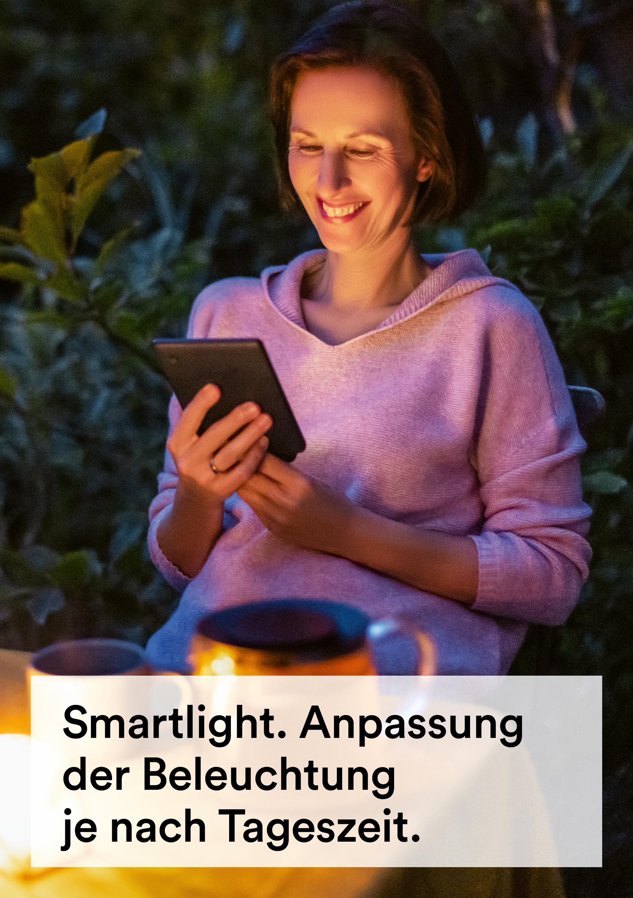 Eine Frau lächelt und betrachtet ein Tablet. Sie trägt einen rosa Pullover. Der Hintergrund ist verschwommen.