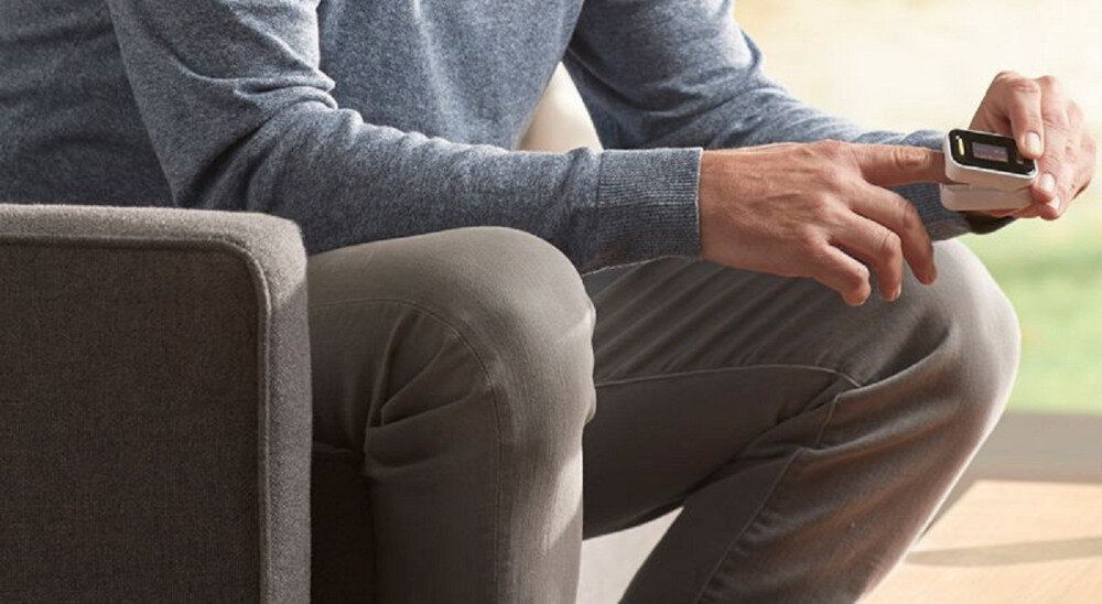 Osoba siedzi, ubrana w szare ubrania, używając pulsoksymetru na palcu. Tło jest rozmyte.