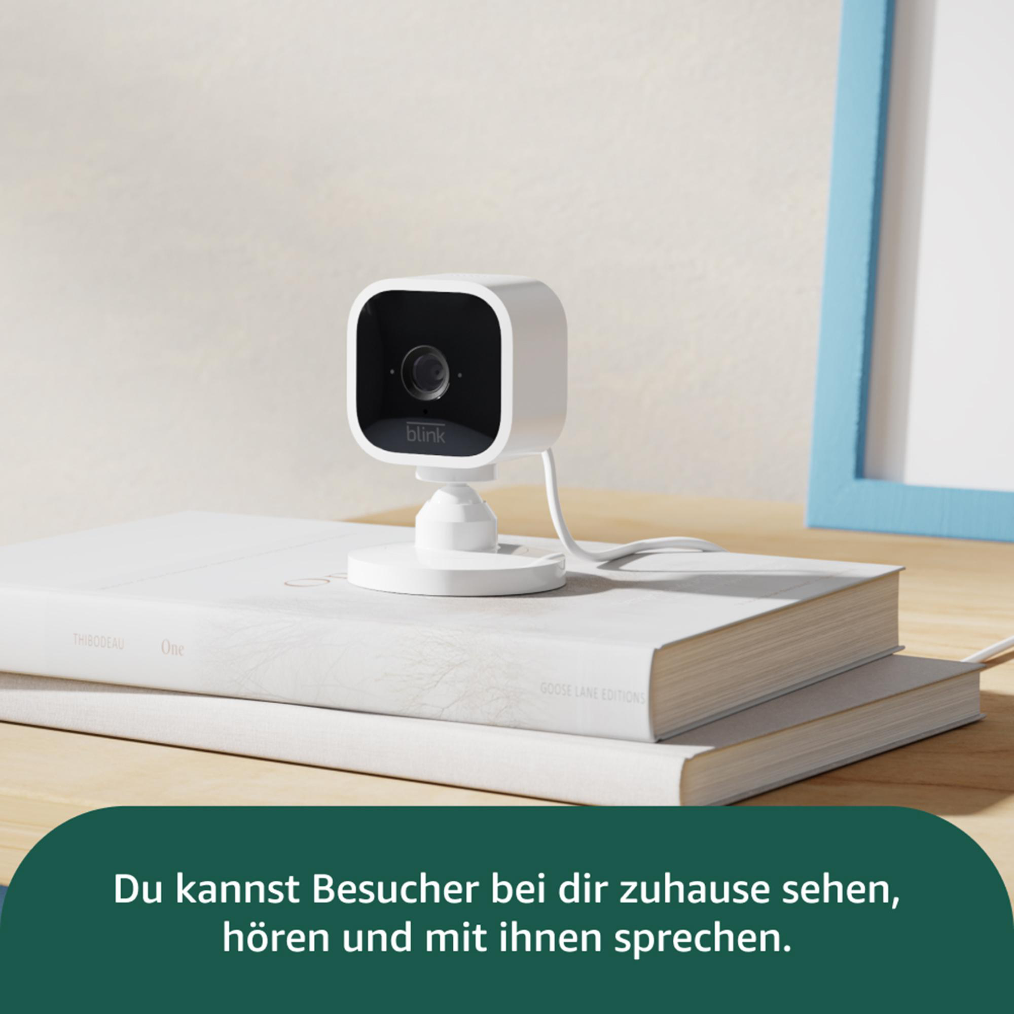 Eine weiße Blink-Sicherheitskamera liegt auf Büchern. Ein blau gerahmtes Foto befindet sich im Hintergrund.