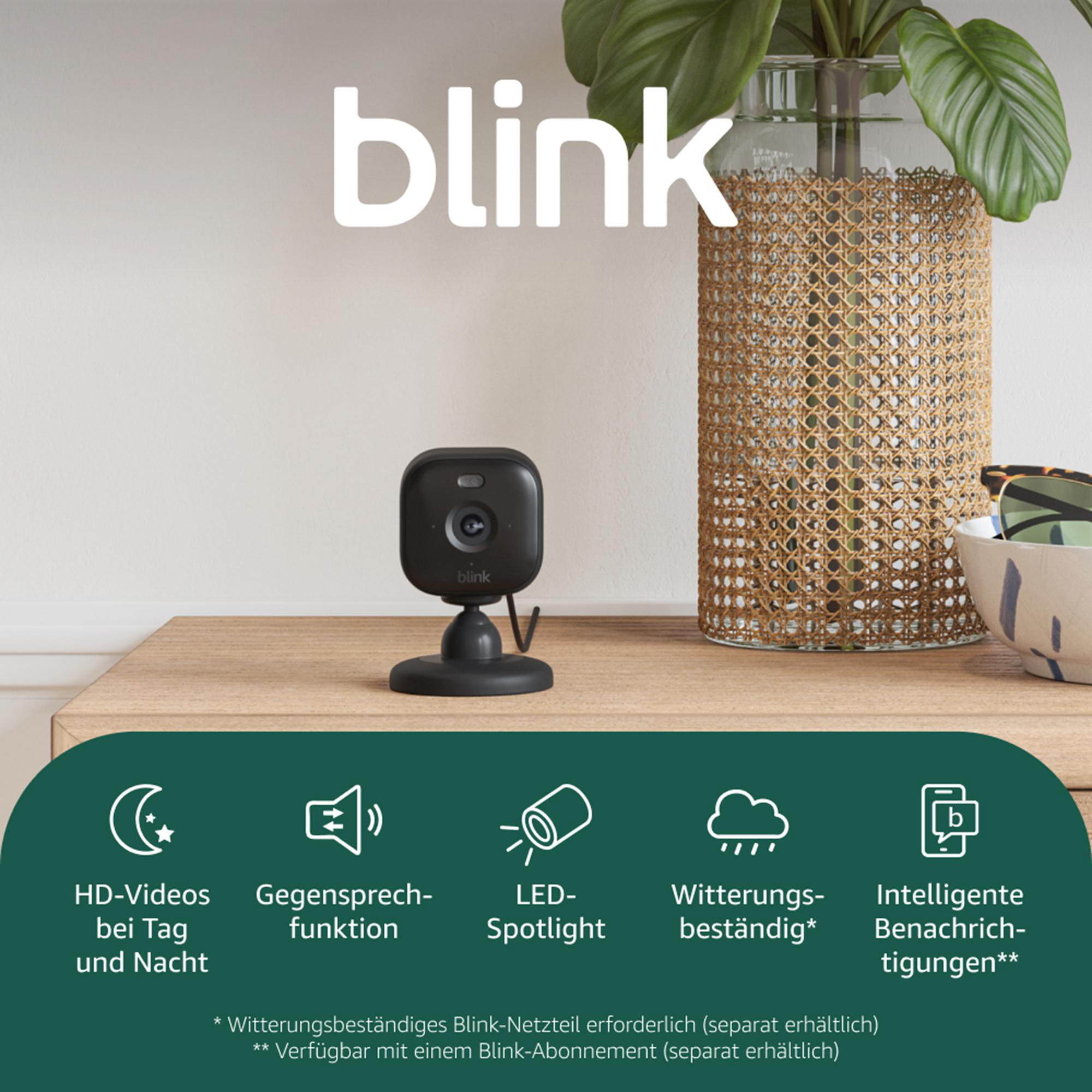 Eine schwarze Blink-Kamera steht auf einer Holzoberfläche. Rechts davon eine Vase mit Pflanzen. Das Wort 'blink' steht oben.
