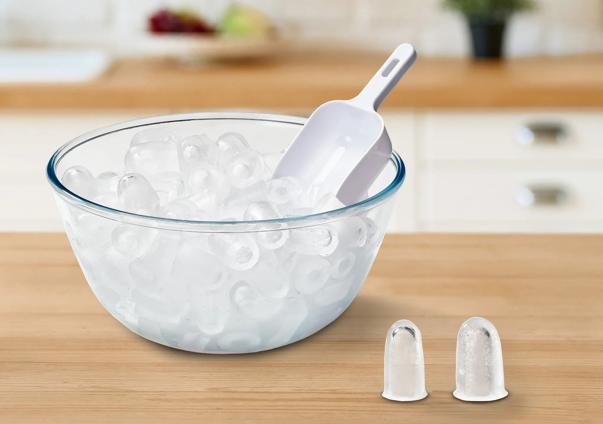 Szklana miska wypełniona kostkami lodu i biała szufelka na drewnianej powierzchni. Dwie foremki do lodu z przodu.