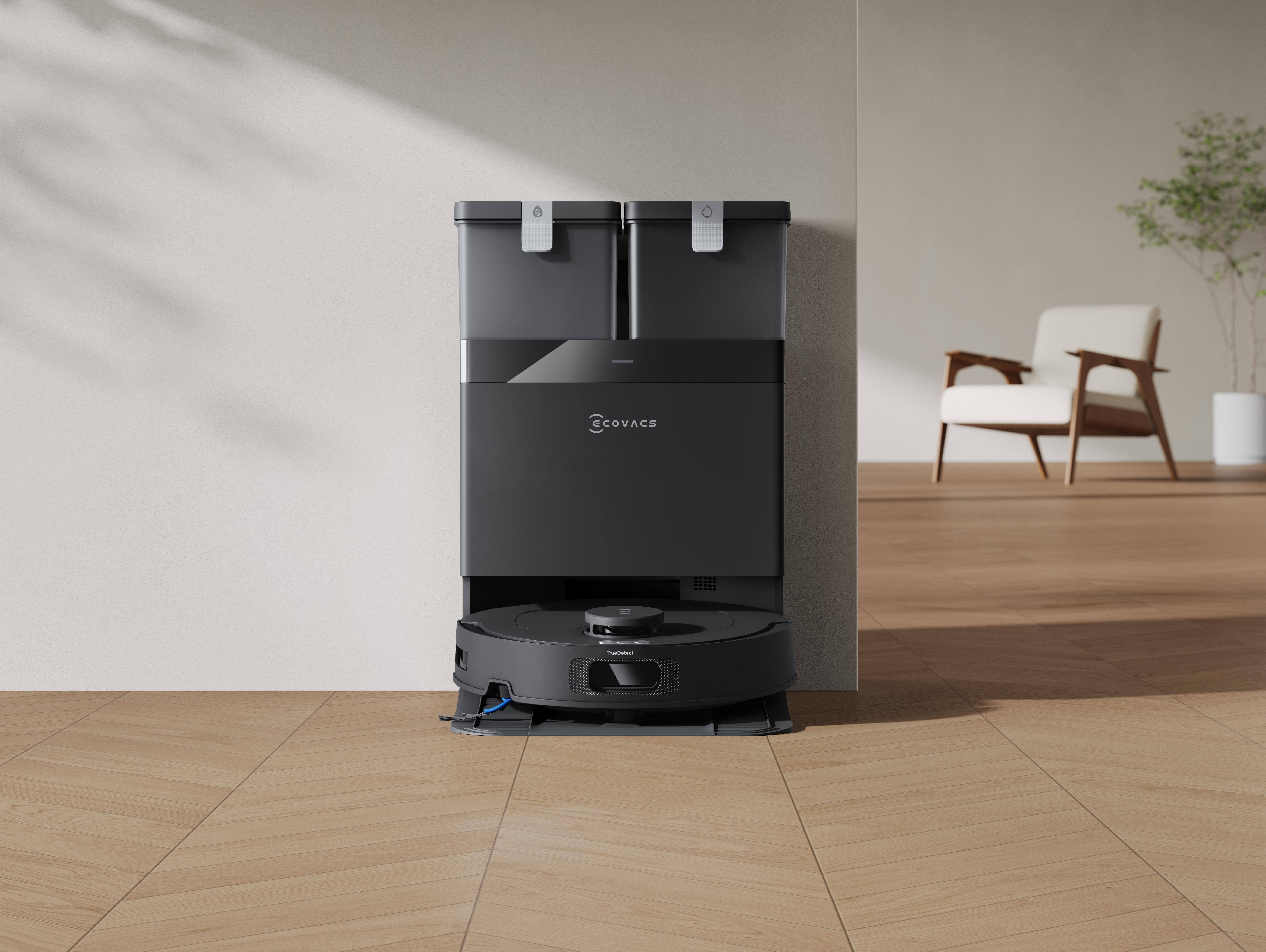 Ein schwarzer Roboterstaubsauger und eine Ladestation stehen auf einem Holzboden. Ein Stuhl und eine Pflanze sind im Hintergrund.