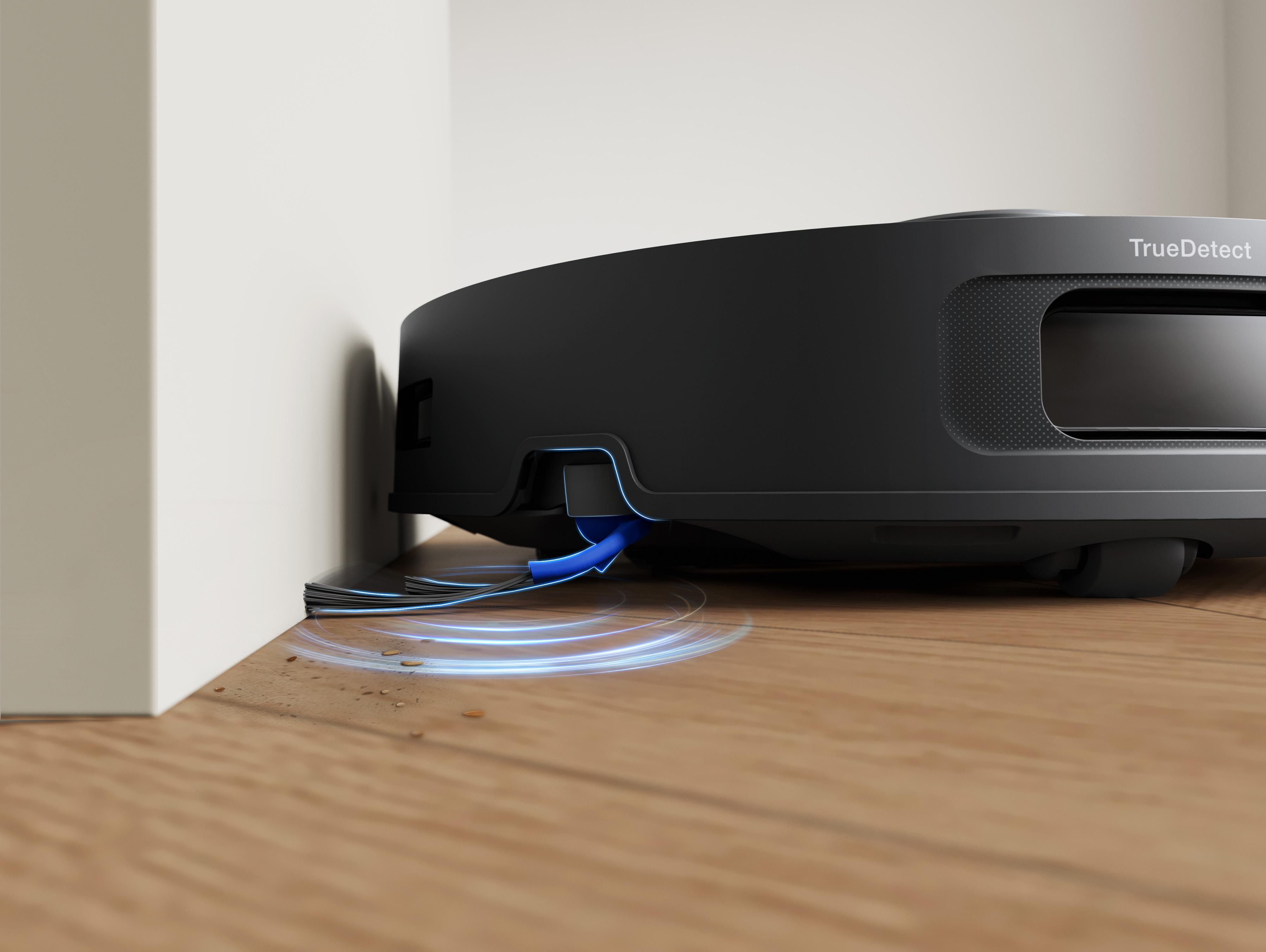 Ein schwarzer Roboter-Staubsauger in der Nähe einer weißen Wand. Er steht auf einem Holzboden mit etwas Schmutz. Der Reiniger hat eine blaue Bürste.