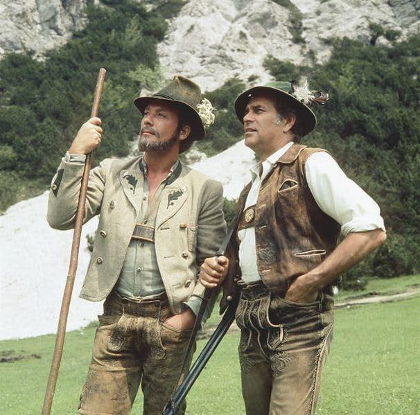 Zwei Männer in traditioneller bayerischer Tracht stehen auf einem grünen Hügel, Berge im Hintergrund. Einer hält einen Stab, der andere ein Gewehr.
