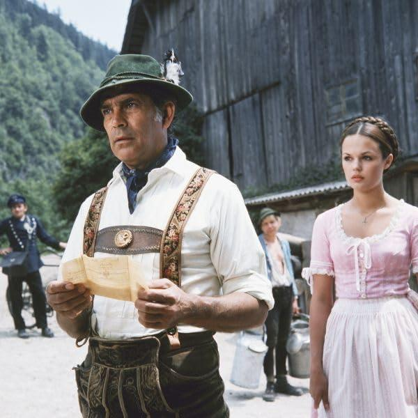 Ein Mann in Hut und Tracht liest ein Papier. Eine Frau steht neben ihm in einem rosa Kleid. Andere sind im Hintergrund.