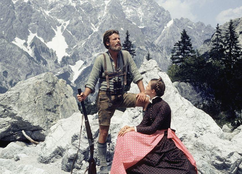 Ein Mann in Lederhosen und eine Frau in einem Kleid posieren auf Felsen, Berge im Hintergrund. Der Mann hält ein Gewehr.
