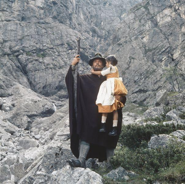 Ein Mann mit Hut und Umhang hält ein Kind. Sie stehen auf Felsen vor einem bergigen Hintergrund und etwas Grün.