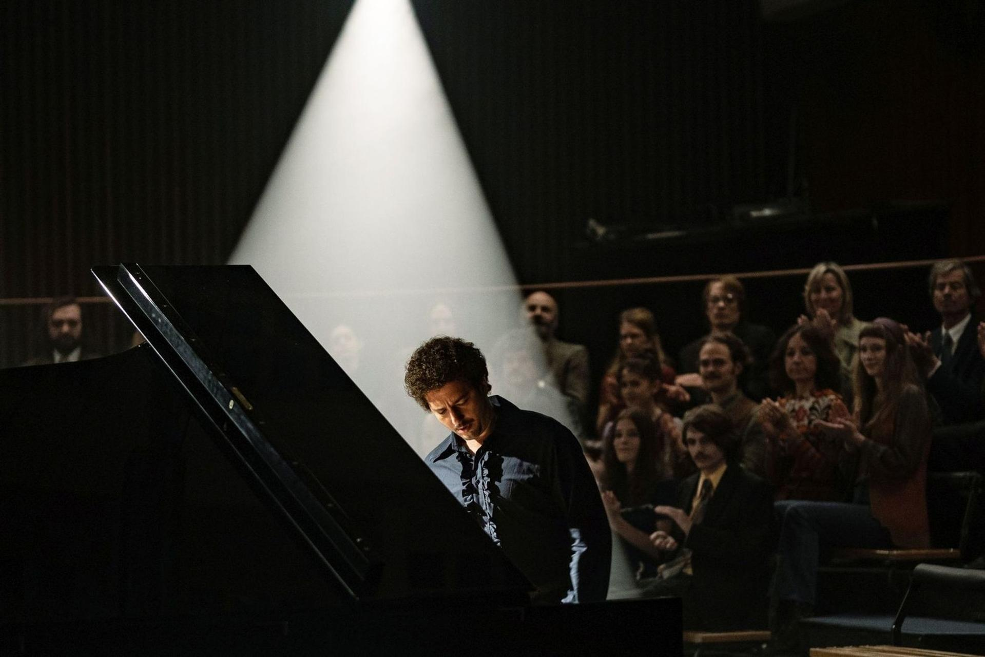 Ein Pianist im Rampenlicht spielt Klavier. Im Hintergrund applaudiert ein Publikum. Die Bühne ist dunkel mit hellem Licht.