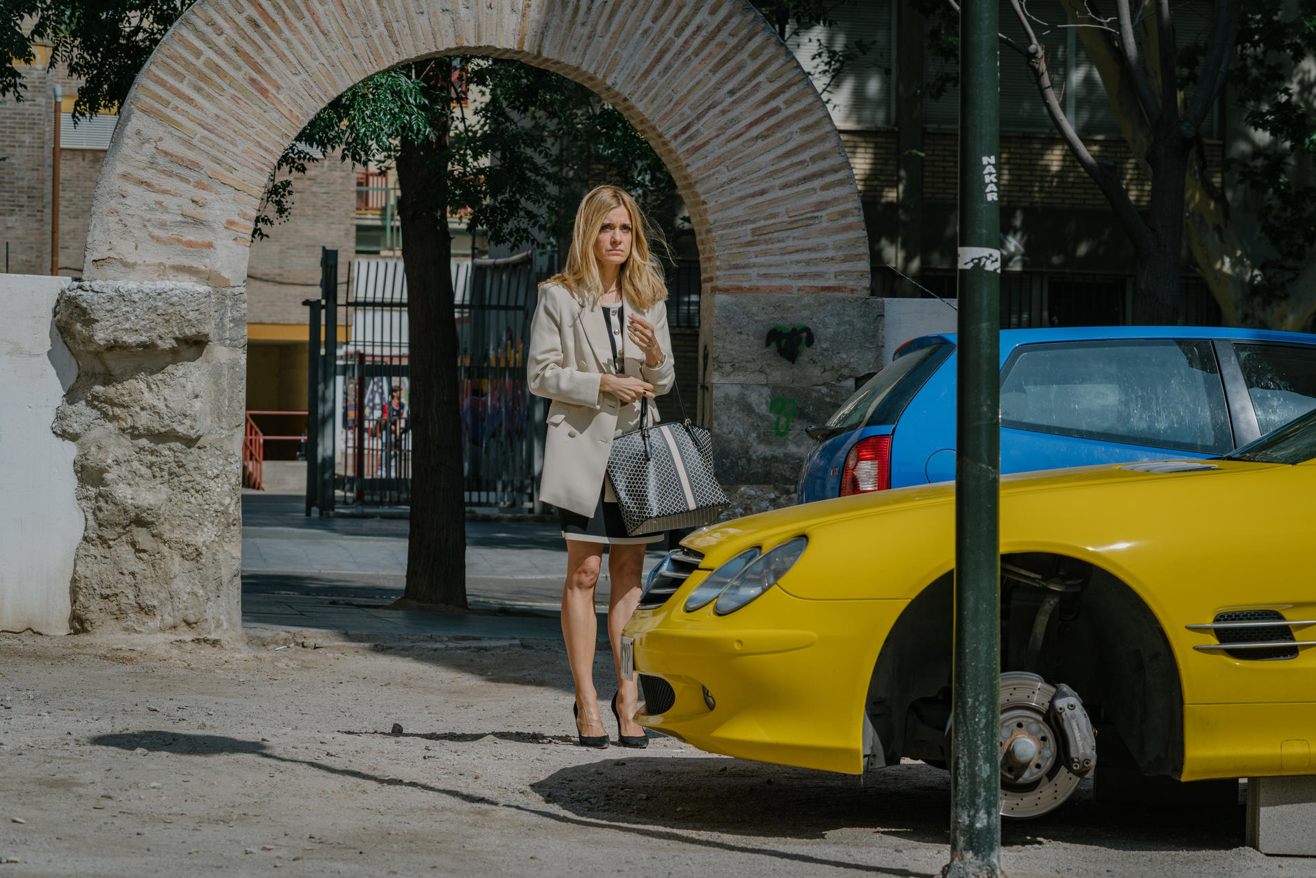Eine Frau in einem beigen Blazer steht in der Nähe eines gelben Autos. Ein Torbogen und ein blaues Auto sind im Hintergrund.