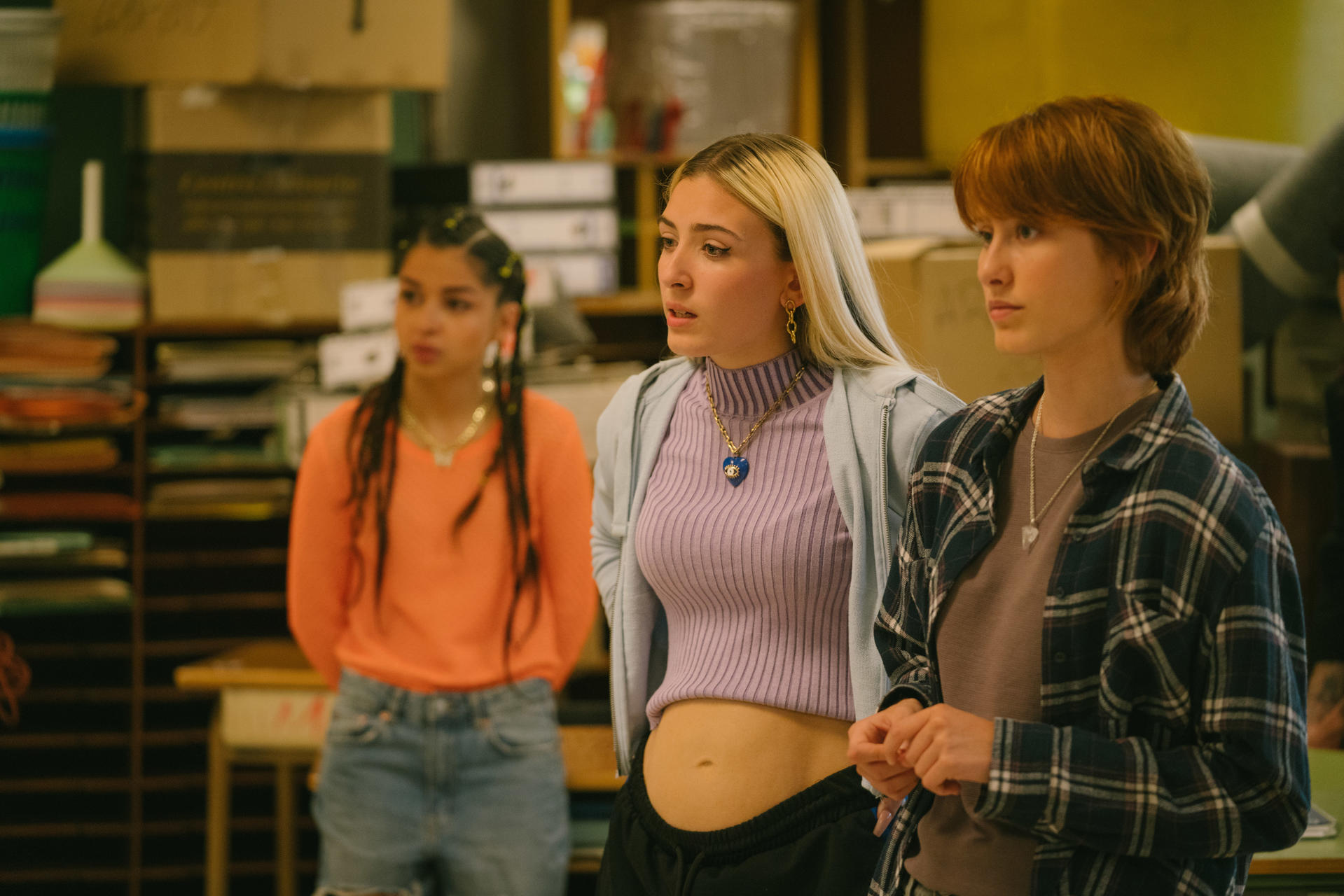 Drei junge Frauen stehen in einem Raum. Eine trägt Orange, eine andere einen lila Pullover und die dritte ein kariertes Hemd. Im Hintergrund sind Regale.