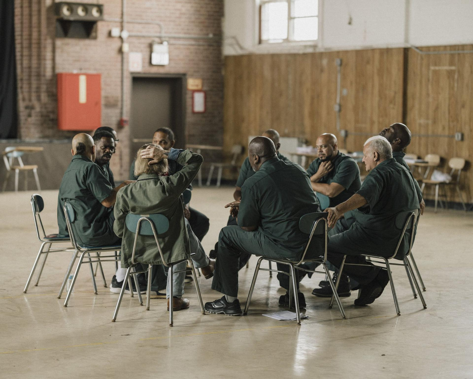 Eine Gruppe von Männern in grünen Uniformen sitzt im Kreis. Sie befinden sich in einem großen Raum. Einige berühren ihre Köpfe.