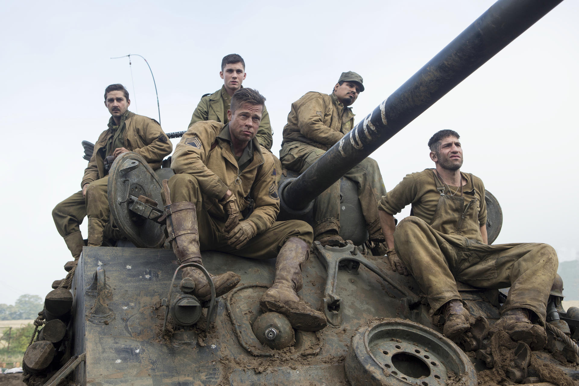 Fünf Soldaten sitzen auf einem schlammigen Panzer. Sie tragen Militäruniformen. Ein Mann in der Mitte schaut in die Kamera.