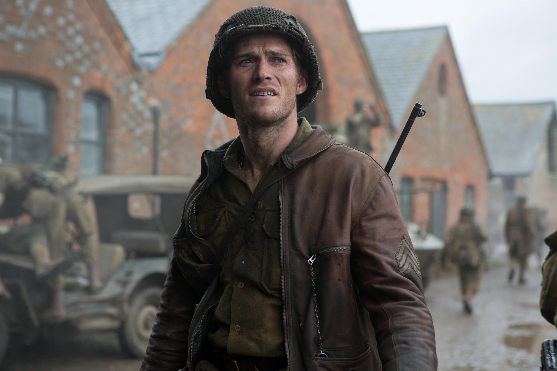 Soldat in WWII-Kleidung, Helm, braune Lederjacke, blickt nach oben, besorgter Gesichtsausdruck. Ziegelgebäude und andere Soldaten im Hintergrund.