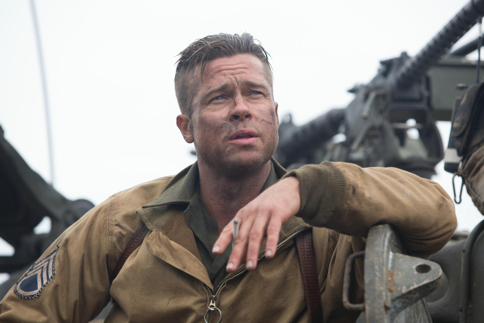 Brad Pitt in WWII-Uniform raucht eine Zigarette. Er blickt nach oben, das Gesicht mit Schmutz verschmiert, hinter ihm ein Maschinengewehr.