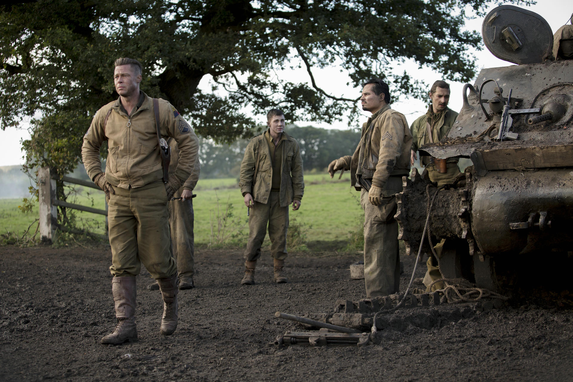 Soldaten in Khaki-Uniformen stehen in der Nähe eines Panzers und eines Zauns. Ein Mann steht mit den Händen in den Hüften. Der Hintergrund zeigt ein Feld.