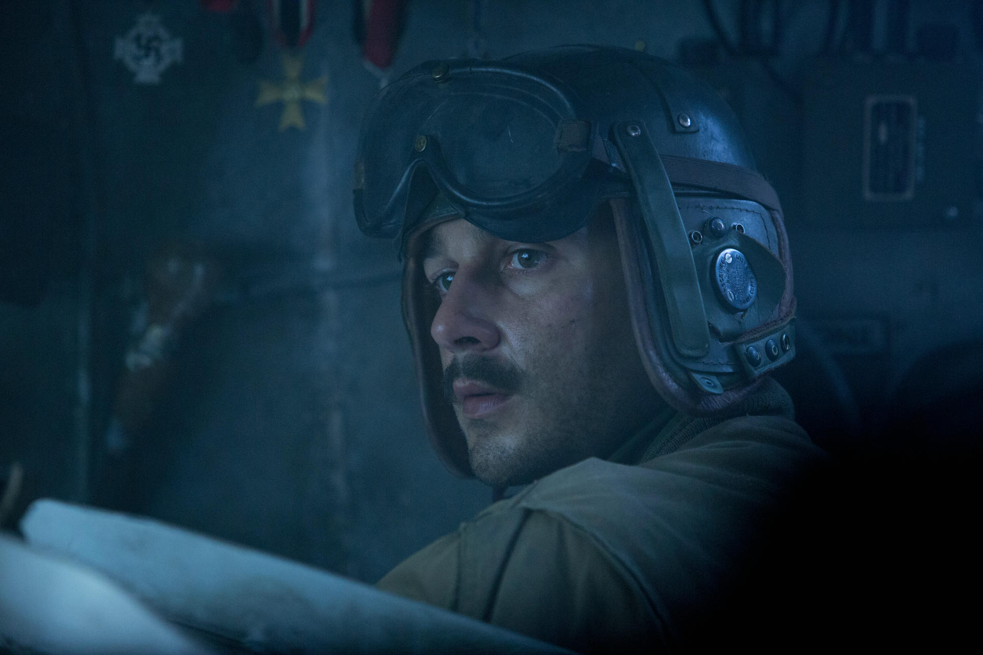 Ein Mann in einem Militärhelm mit Schutzbrille. Er hat einen Schnurrbart und blickt zur Seite. Der Hintergrund ist dunkel.