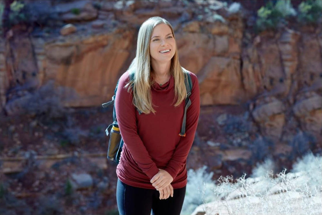 Frau lächelt, trägt bordeauxfarbenes Hemd und Rucksack. Steht in einem Canyon mit Felsformationen im Hintergrund.
