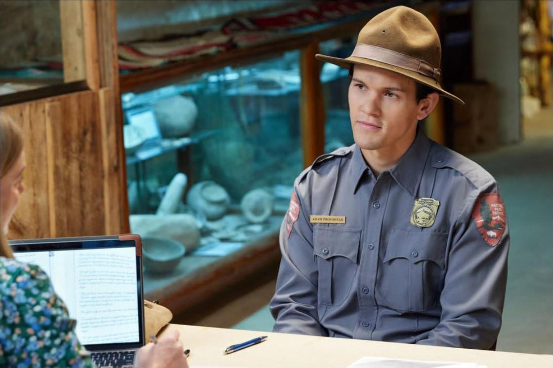 Ein Parkranger in Uniform und Hut blickt eine Frau an. Vor der Frau steht ein Laptop. Im Hintergrund sind Artefakte ausgestellt.