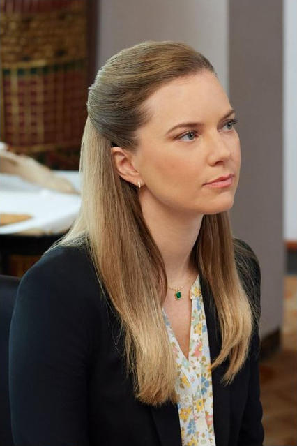 Eine Frau mit blondem Haar und einem schwarzen Blazer blickt zur Seite. Sie trägt ein Blumenhemd und eine grüne Halskette.
