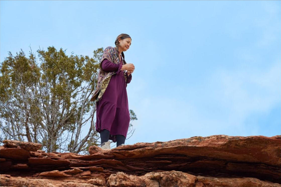 Frau im lila Kleid steht auf einem roten Felsen, mit grünen Bäumen und blauem Himmel im Hintergrund.
