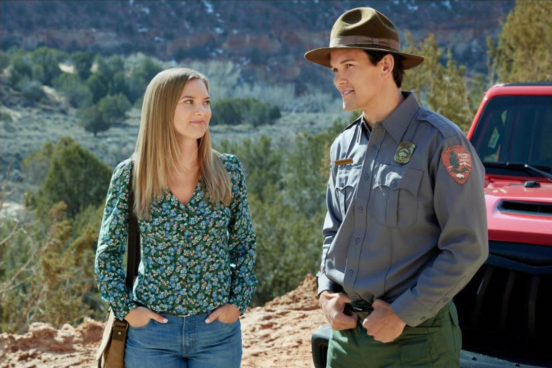 Eine Frau und ein Park Ranger stehen sich gegenüber. Die Frau trägt ein Blumenhemd. Der Ranger trägt eine Uniform und einen Hut. Ein roter Jeep ist hinter ihnen.