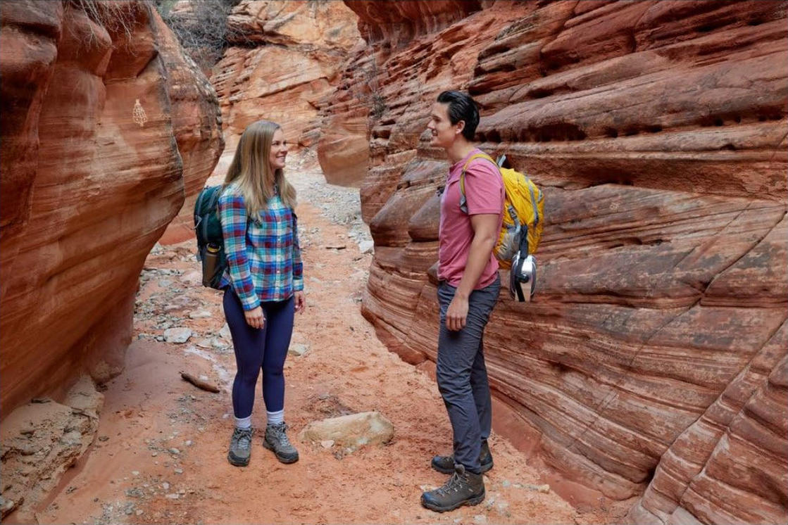 Zwei Wanderer stehen in einem schmalen Canyon. Rote Felswände umgeben sie. Einer trägt ein kariertes Hemd, der andere ein rosa Hemd. Beide haben Rucksäcke.