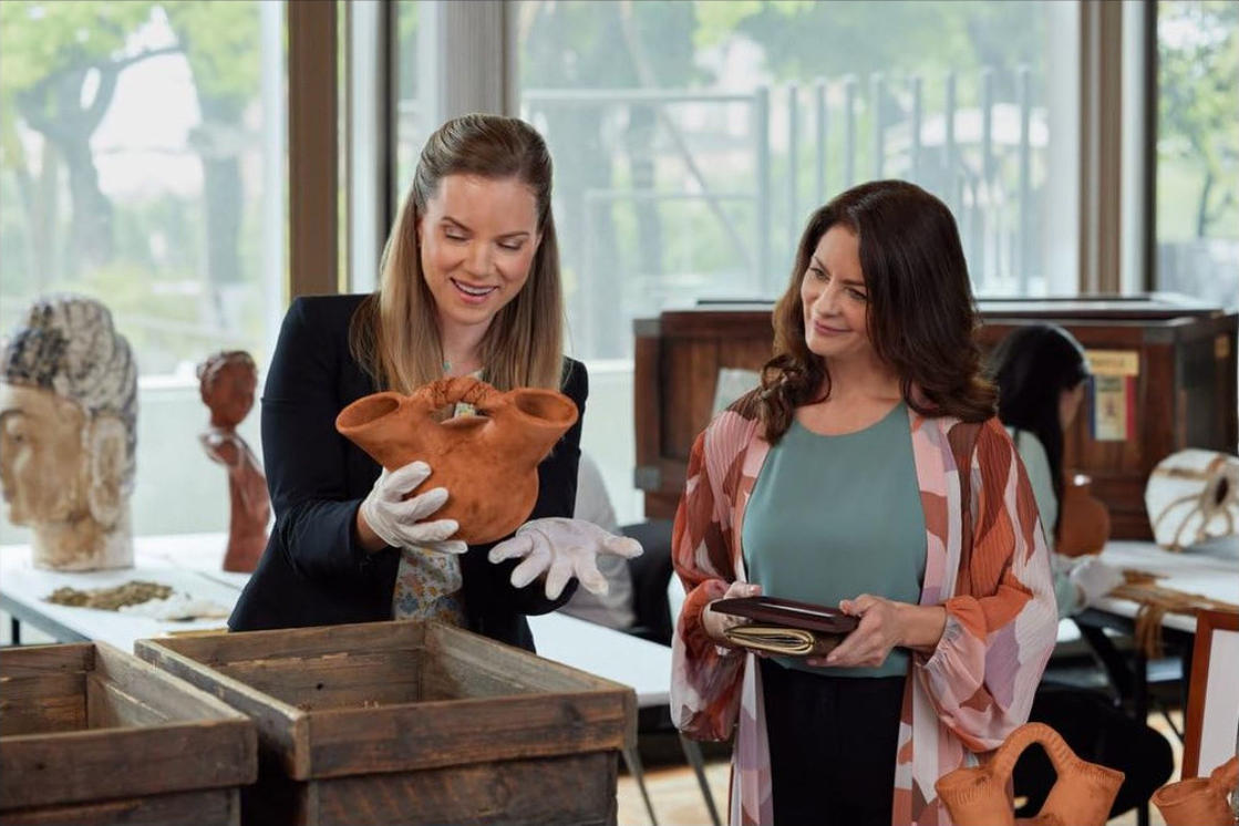 Zwei Frauen begutachten ein Tongefäß in einem Museum. Eine hält es mit Handschuhen, die andere hält eine Brieftasche. Statuen werden ausgestellt.