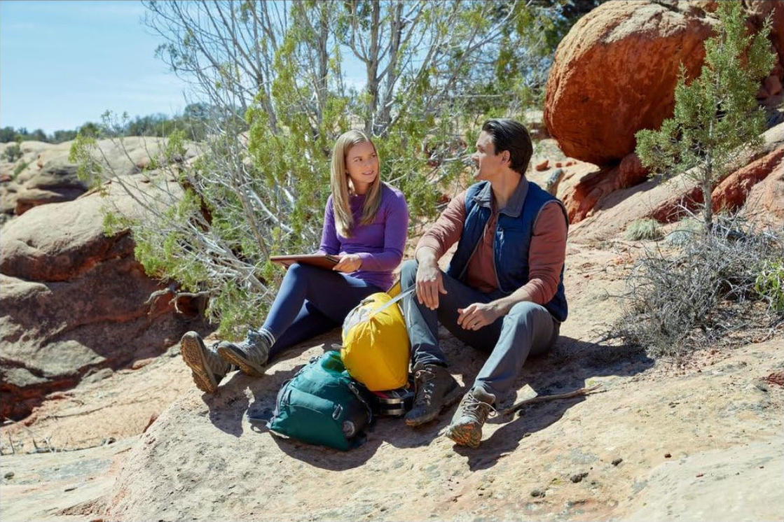 Ein Mann und eine Frau sitzen auf Felsen und unterhalten sich. Rucksäcke und eine gelbe Tasche liegen neben ihnen. Bäume und Felsen umgeben sie.