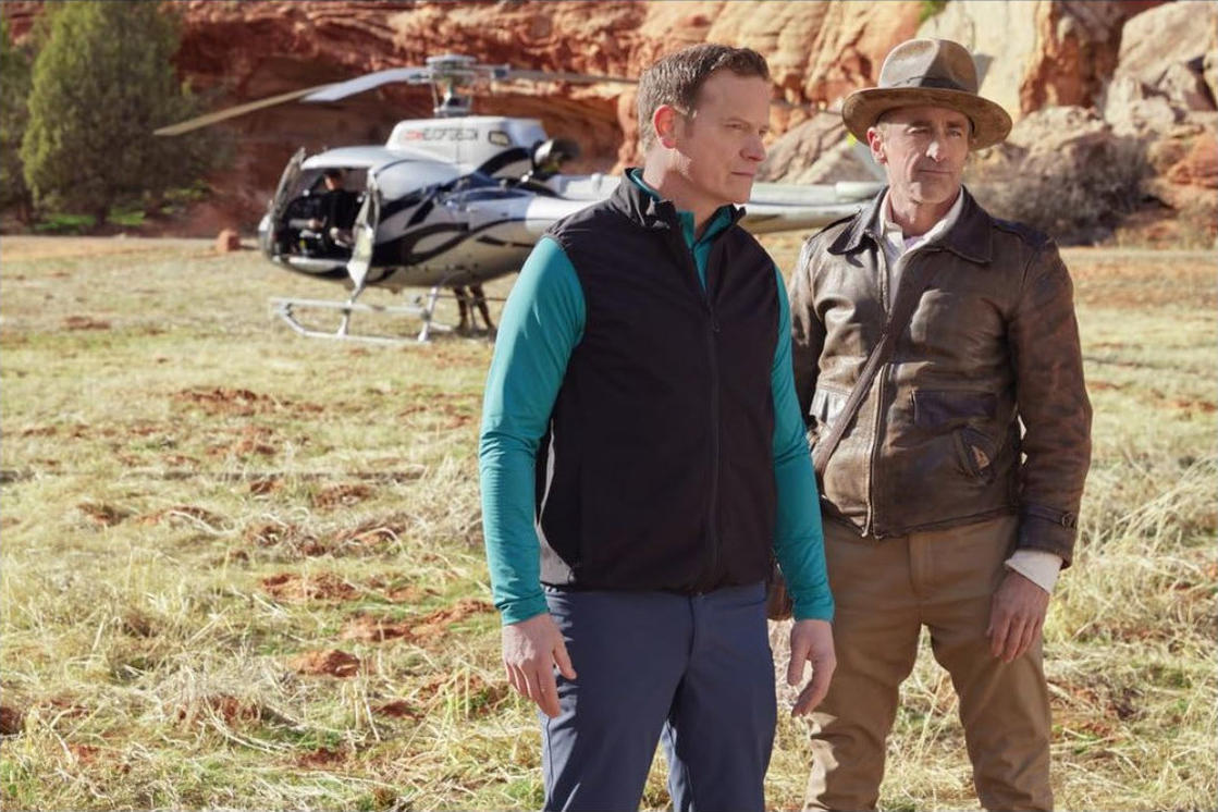 Zwei Männer stehen in der Nähe eines Hubschraubers auf einem Feld. Einer trägt eine Weste, der andere eine Lederjacke und einen Hut. Roter Felsen im Hintergrund.