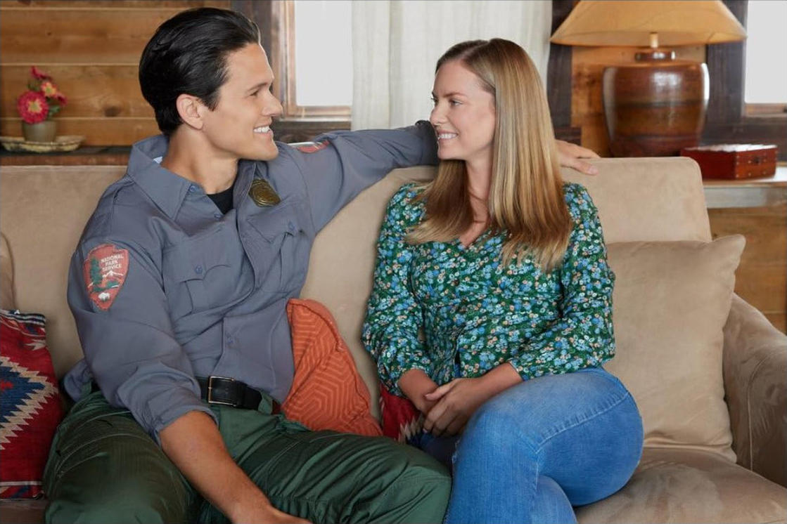 Ein Mann in Uniform und eine Frau sitzen auf einem Sofa und lächeln sich an. Sie sind drinnen, mit einer Lampe und Blumen im Hintergrund.