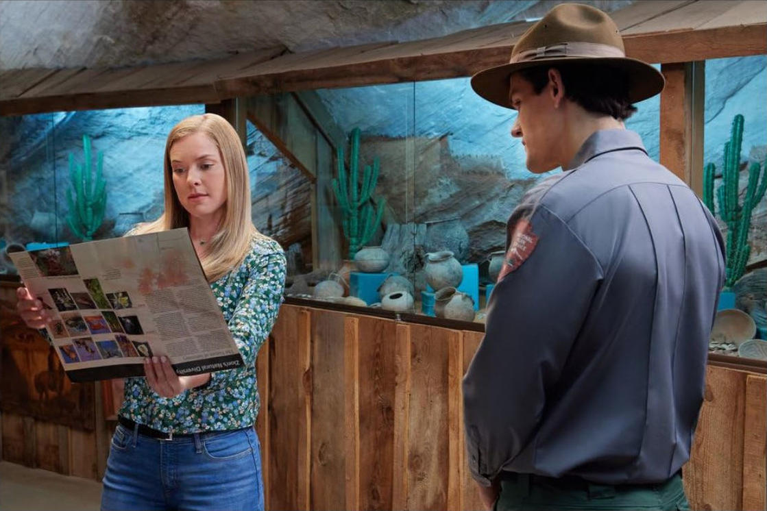 Eine Frau liest eine Zeitung. Ein Park Ranger steht in der Nähe. Im Hintergrund sind Kakteen, Töpferwaren und eine Gesteinsausstellung zu sehen.