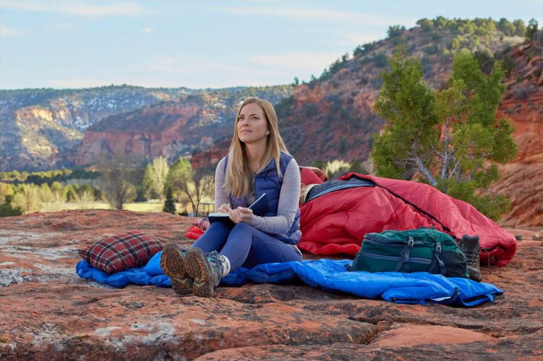 Frau im Camping-Setting. Sitzt auf einem Felsen mit Notizbuch. Rote Berge im Hintergrund, roter Schlafsack, grüne Tasche und Stiefel in der Nähe.