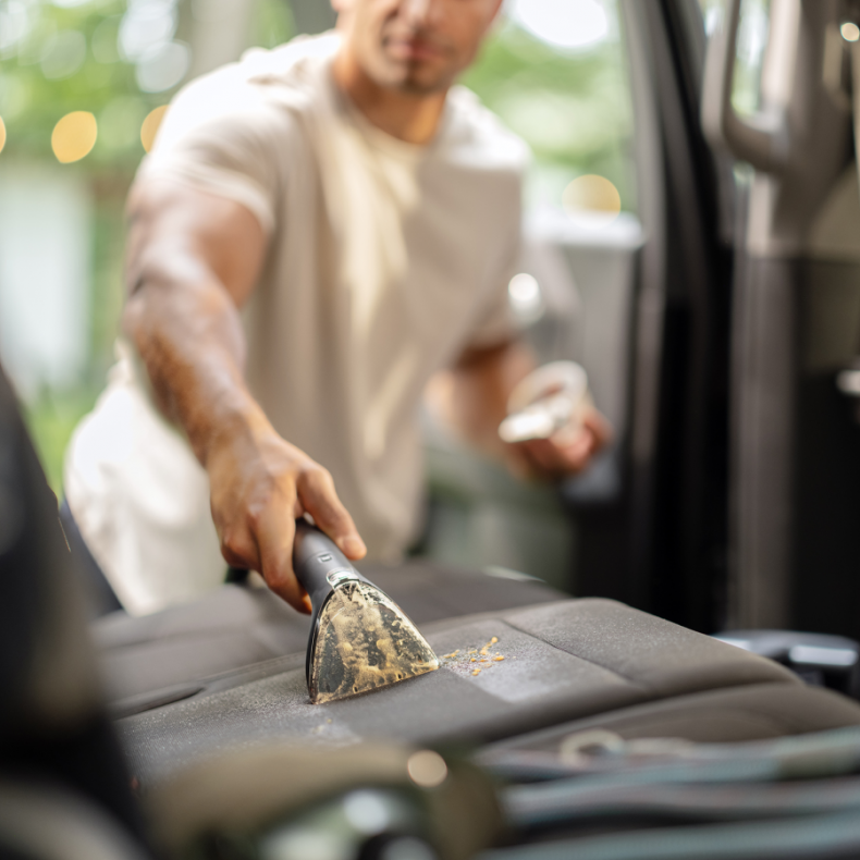 Mężczyzna odkurza fotel samochodowy odkurzaczem ręcznym. Siedzenie jest ciemnoszare, a głowica odkurza kurz.