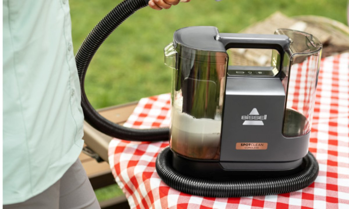 Osoba trzyma wąż podłączony do odkurzacza Bissell SpotClean na czerwono-szachowym stole piknikowym. Zielone tło.