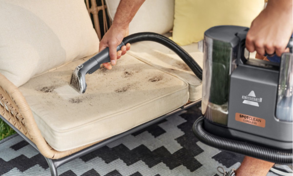 Osoba czyści brudną poduszkę zewnętrzną za pomocą Bissell SpotClean Cordless. Widoczna jest maszyna i wąż.