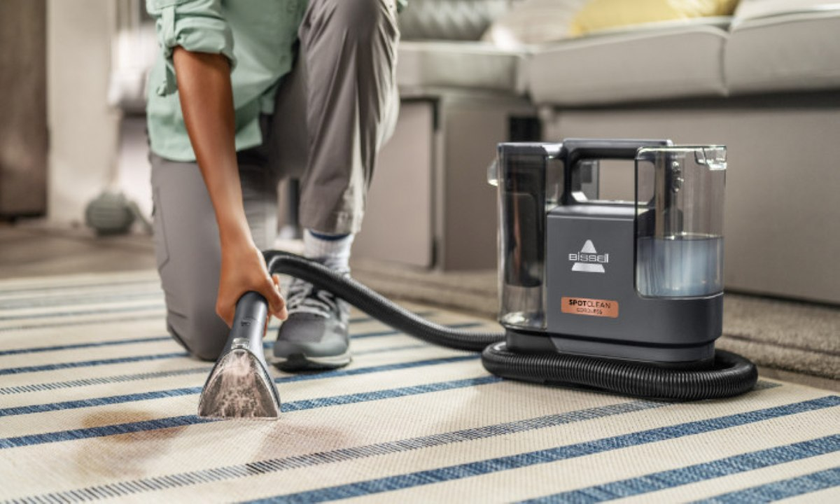 Osoba klęczy i używa odkurzacza Bissell SpotClean na pasiastym dywanie. Odkurzacz czyści plamę.