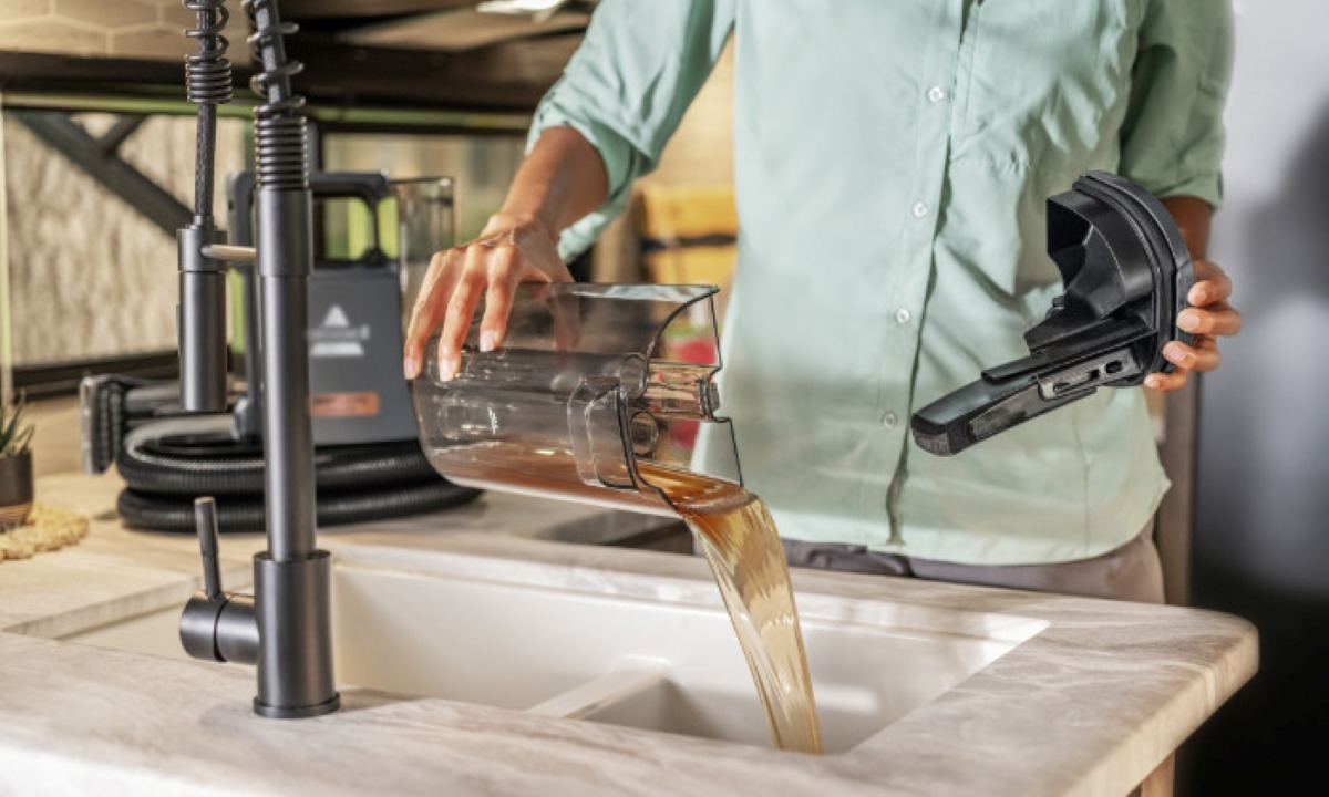 Osoba wlewa płyn z przezroczystego pojemnika do zlewu. W drugiej ręce trzyma czarną część urządzenia.