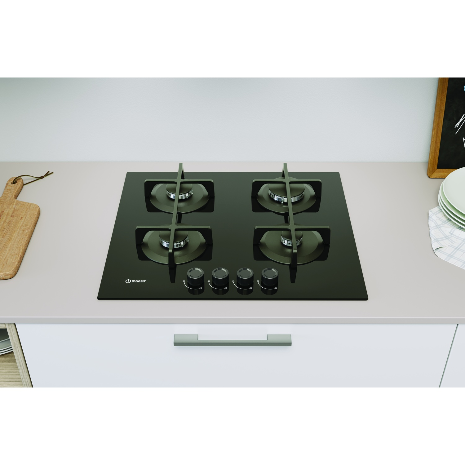 Un piano cottura a gas Indesit in vetro nero con quattro fuochi è su un piano di lavoro grigio chiaro. Sono visibili anche un tagliere in legno e dei piatti.
