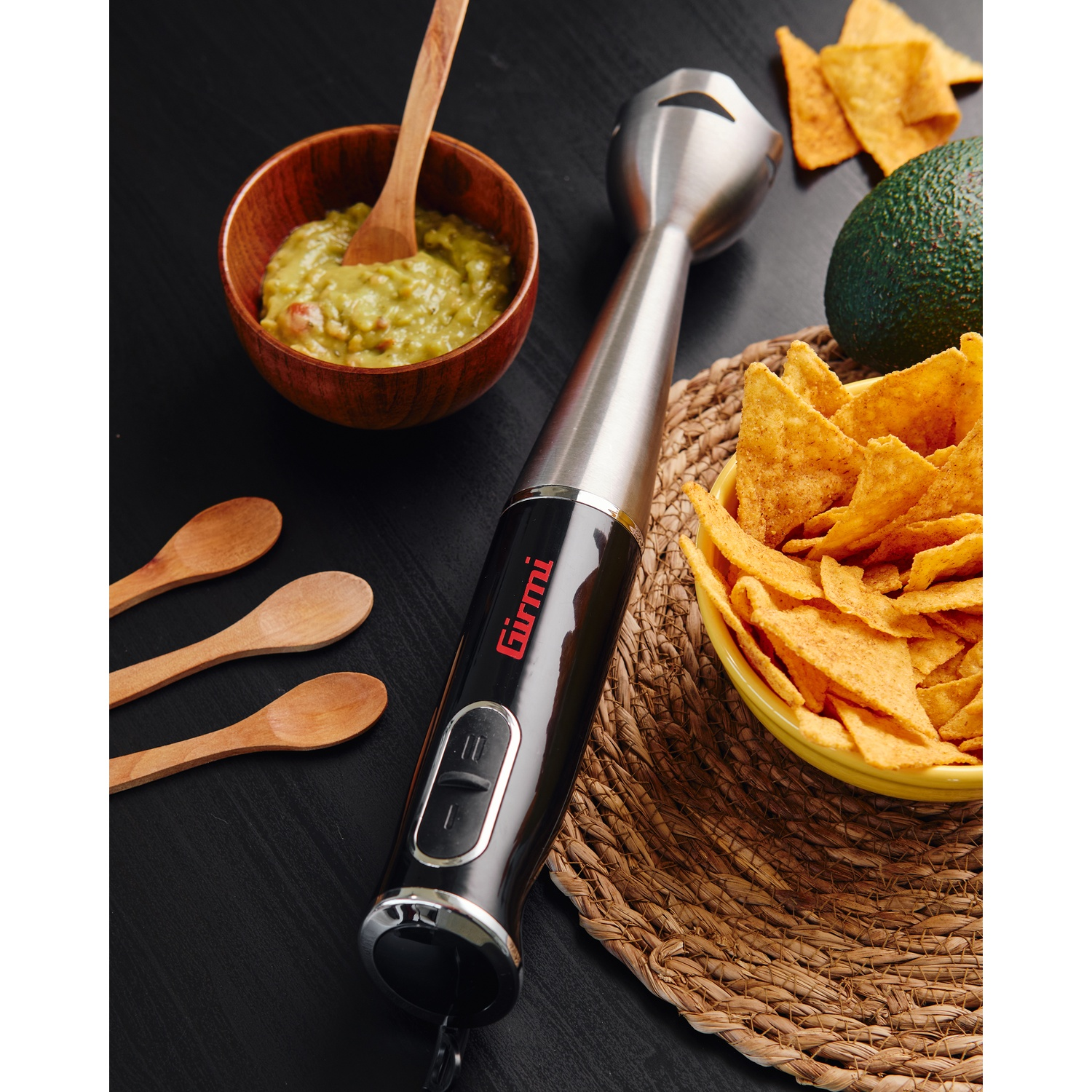 Un frullatore a immersione nero e argento con guacamole, avocado, nachos e cucchiai di legno su una superficie nera.