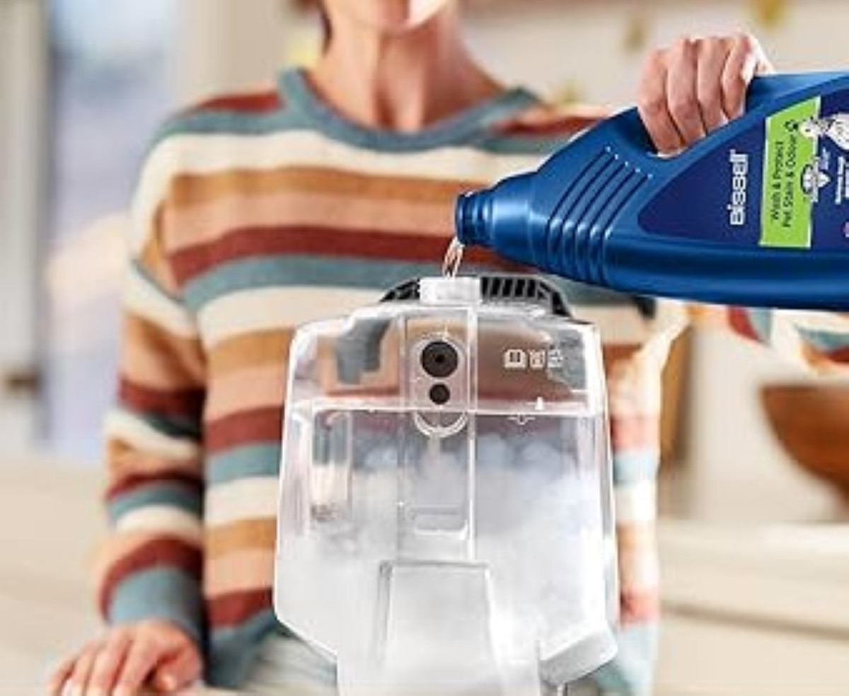 Osoba wlewa płyn z niebieskiej butelki do przezroczystego urządzenia czyszczącego. Ma na sobie sweter w paski.