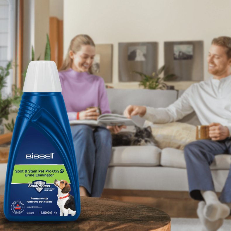 Niebieska butelka Bissell, odplamiacz, stoi na stole. Para relaksuje się na kanapie w tle, z psem.