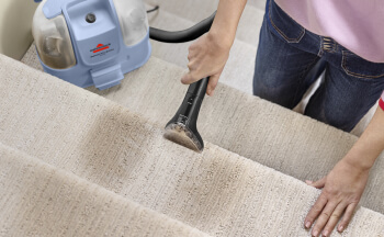 Osoba odkurza schody pokryte wykładziną za pomocą niebiesko-białego urządzenia czyszczącego. Schody są jasnobeżowe.
