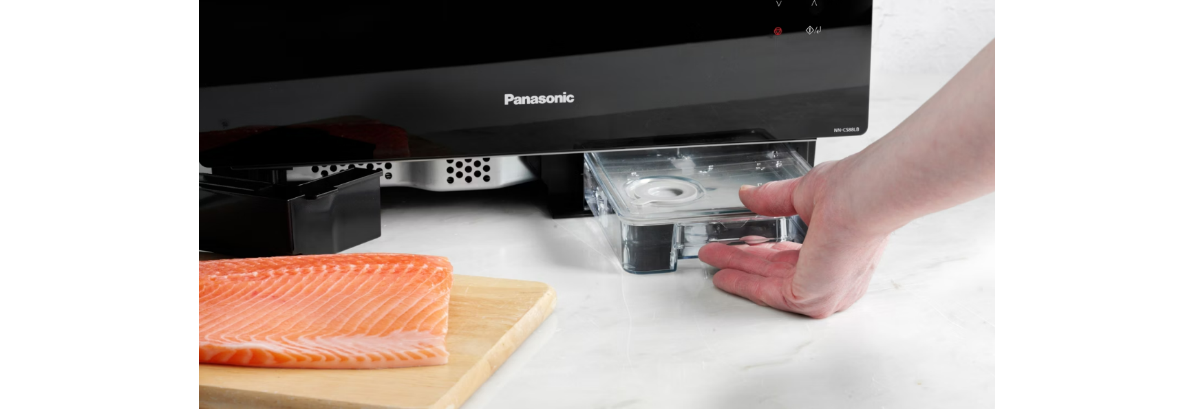 Ręka wyciąga przezroczysty pojemnik z czarnego urządzenia. Łosoś leży na desce do krojenia. Białe tło.