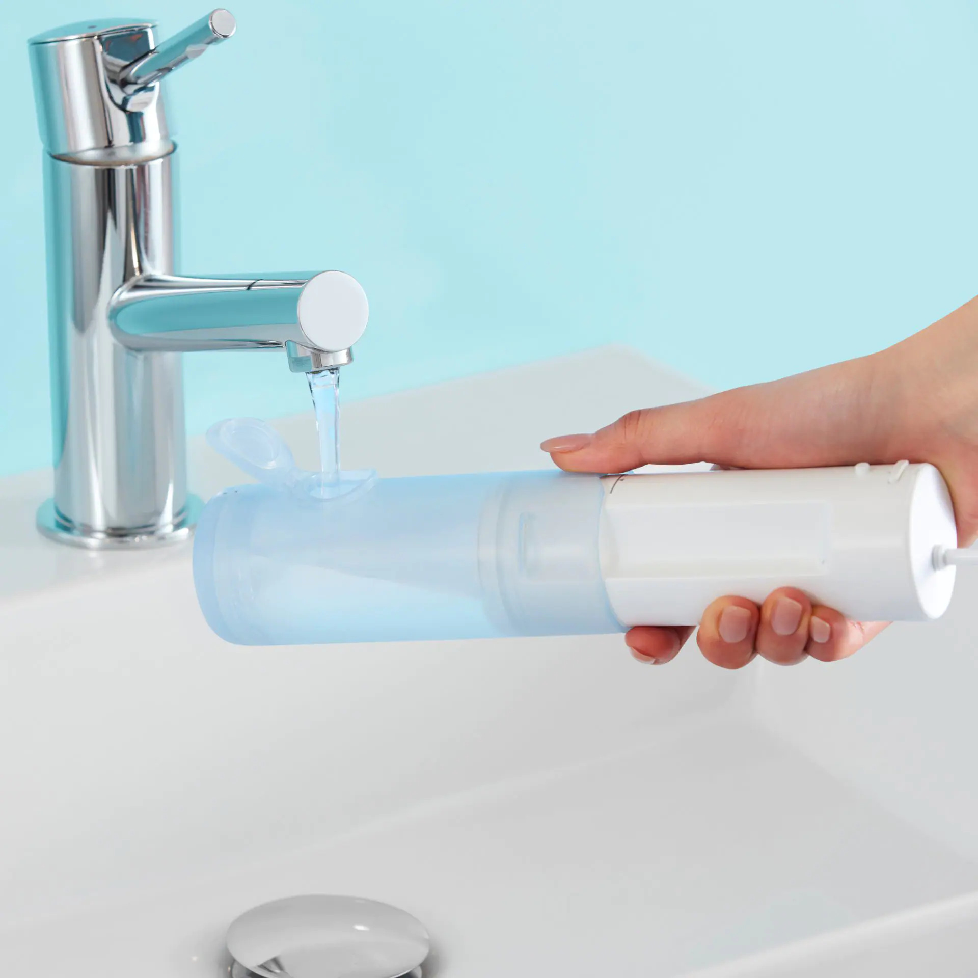 Osoba napełnia niebieski pojemnik wodą z chromowanej baterii nad białą umywalką. W tle znajduje się jasnoniebieska ściana.