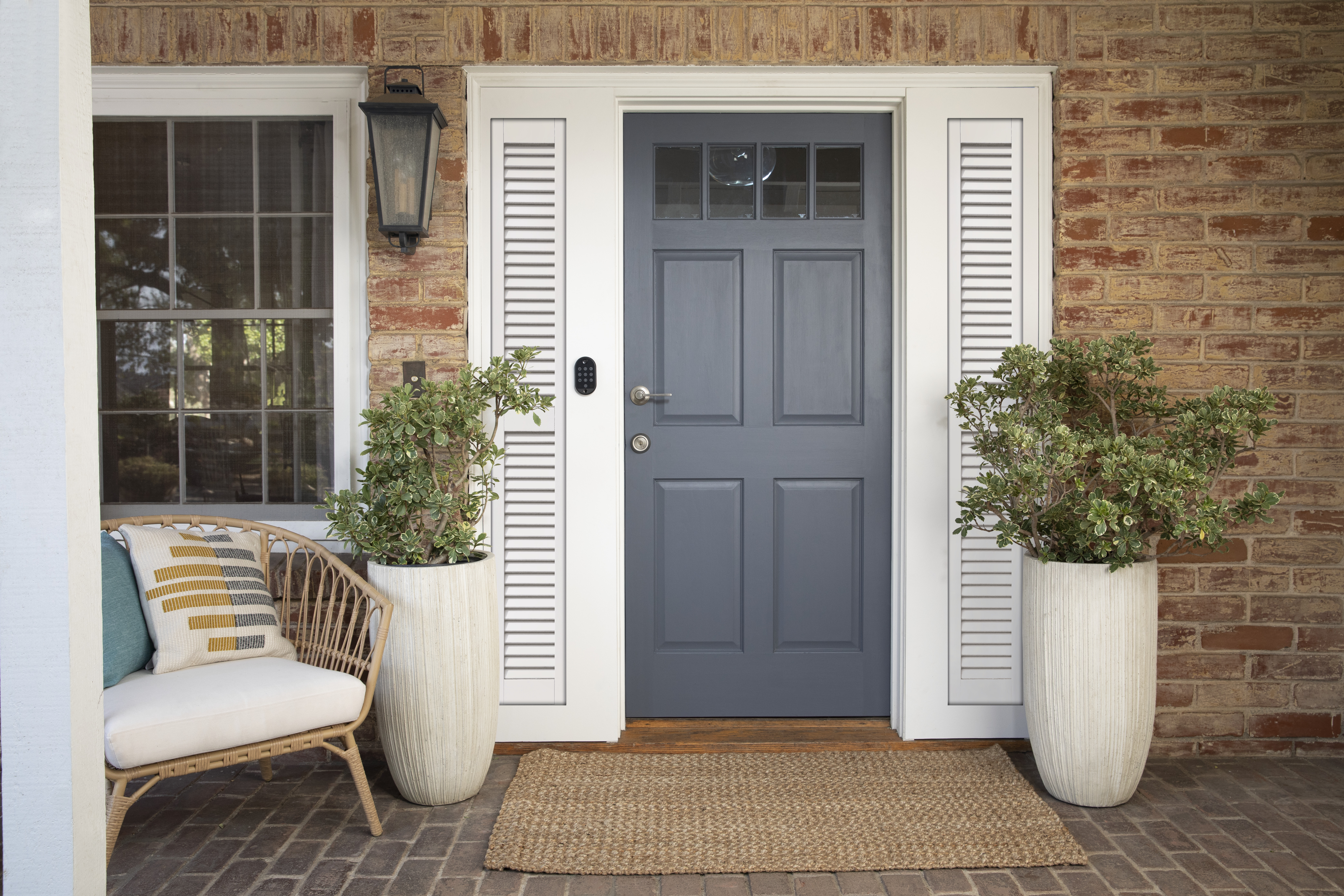 Zentral ist eine blaue Tür, flankiert von weißen Säulen. Links und rechts befinden sich ein Stuhl und Pflanzen, sowie eine Ziegelmauer.