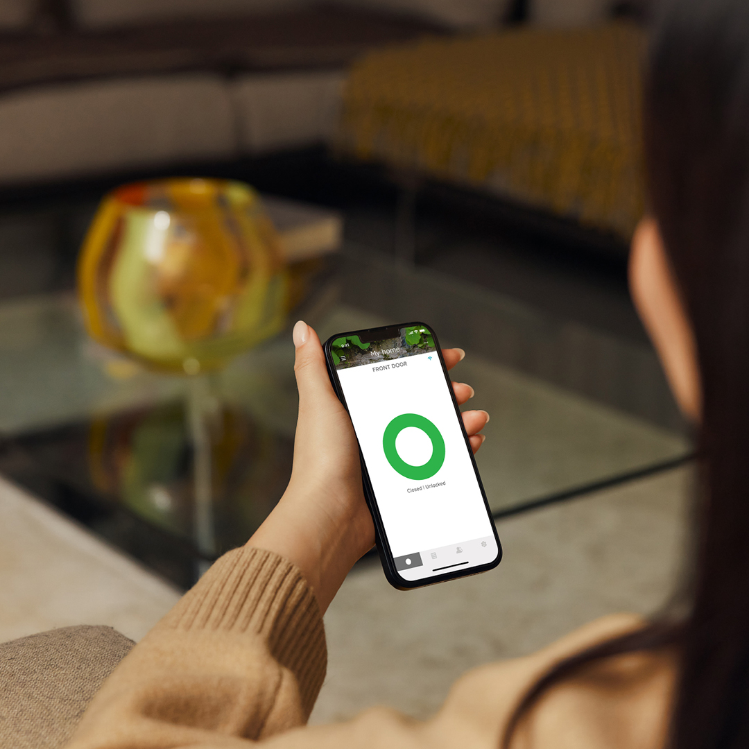 Eine Frau hält ein Telefon mit einer Smart-Lock-Oberfläche. Der Bildschirm zeigt einen grünen Kreis, der anzeigt, dass die Haustür geschlossen und entriegelt ist.